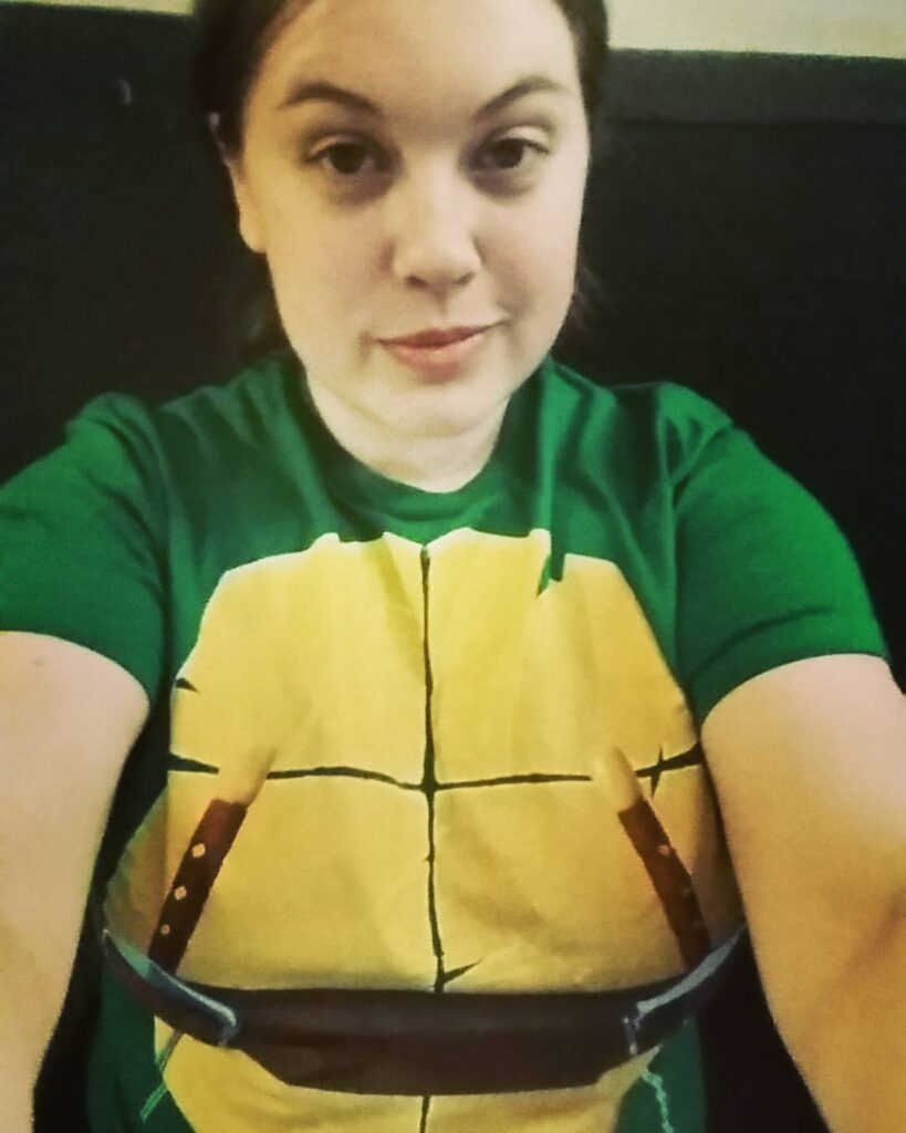 Photo is a selfie of Kristin wearing a Teenage Mutant Ninja Turtle shirt